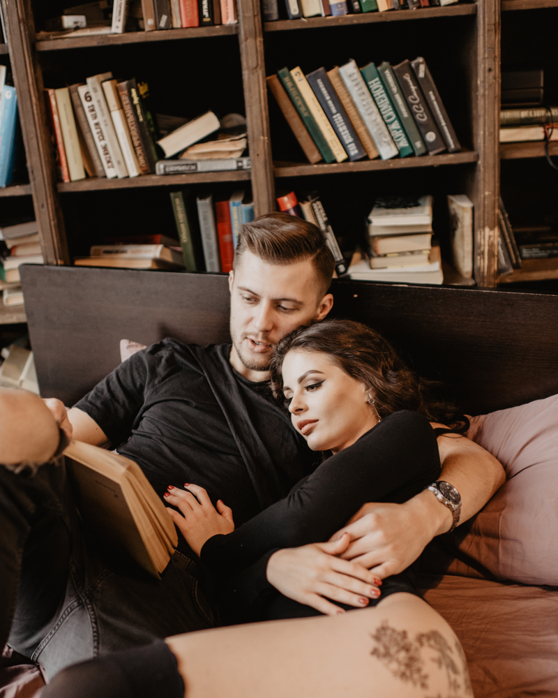 Young,Smiling,Couple,Reading,Book,Sitting,On,Bed,In,Bedroom.