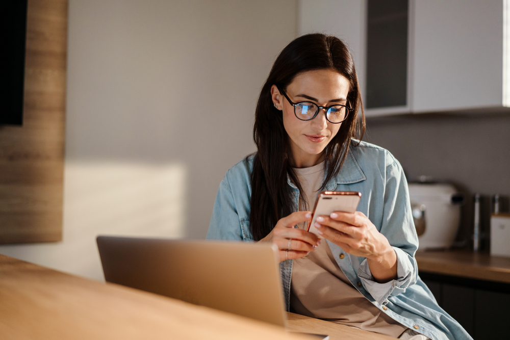 Serious,Charming,Woman,Using,Smartphone,While,Working,With,Laptop,At
