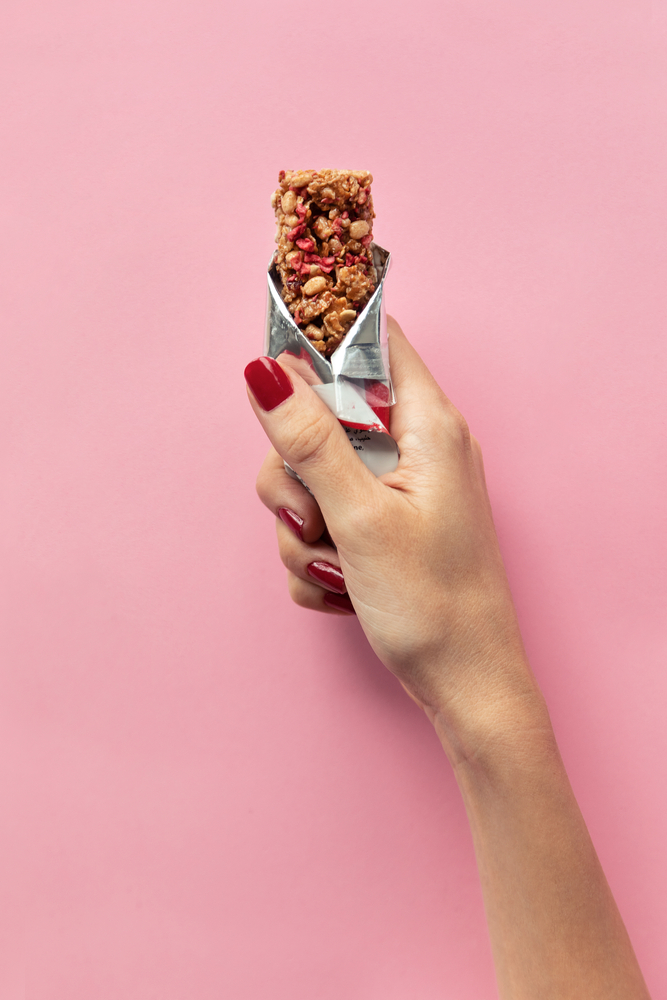 Close-up,Of,Young,Woman,Hand,Holding,Cereal,Bar,On,Pastel