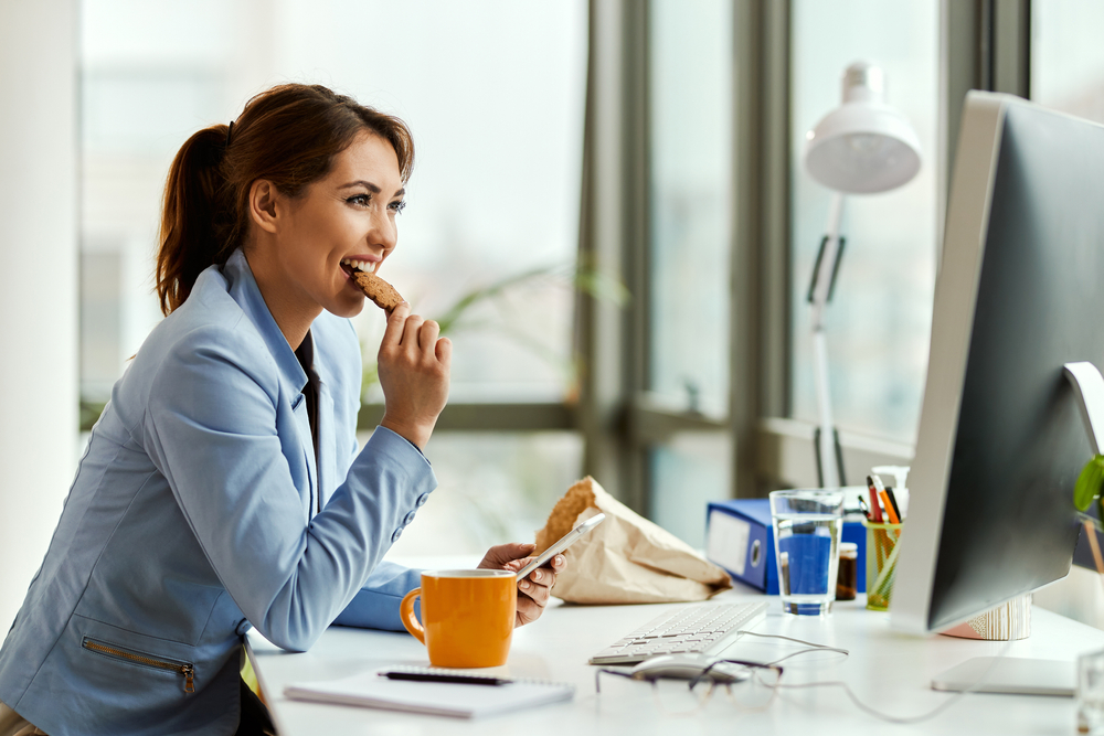 Young,Businesswoman,Using,Cell,Phone,While,Eating,A,Cookie,At