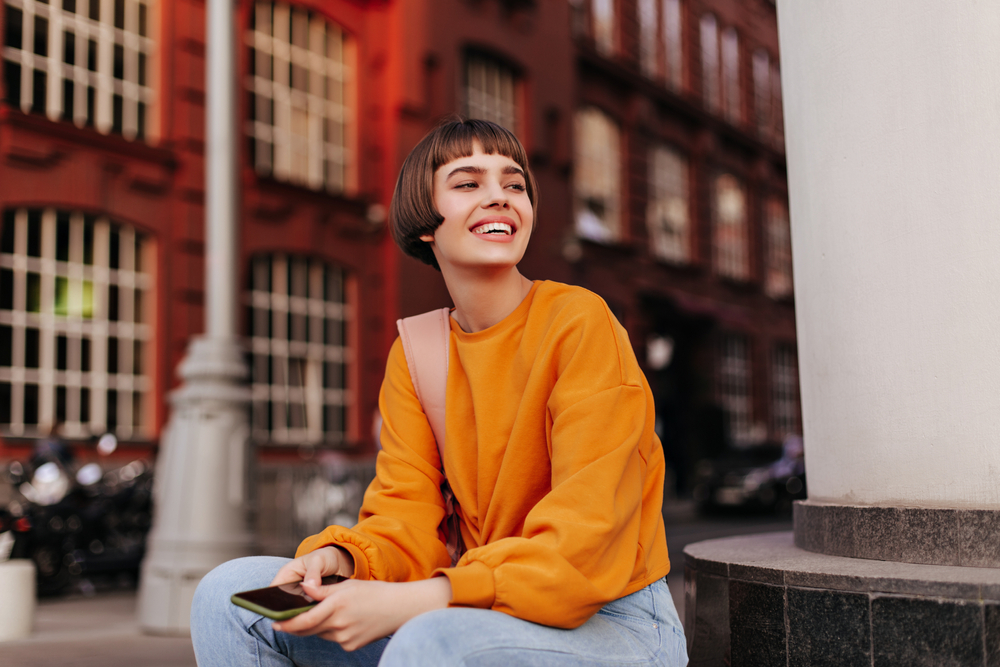 Good-humored,Woman,Sits,Outside.,Short-haired,Girl,In,Orange,Sweater,Holds