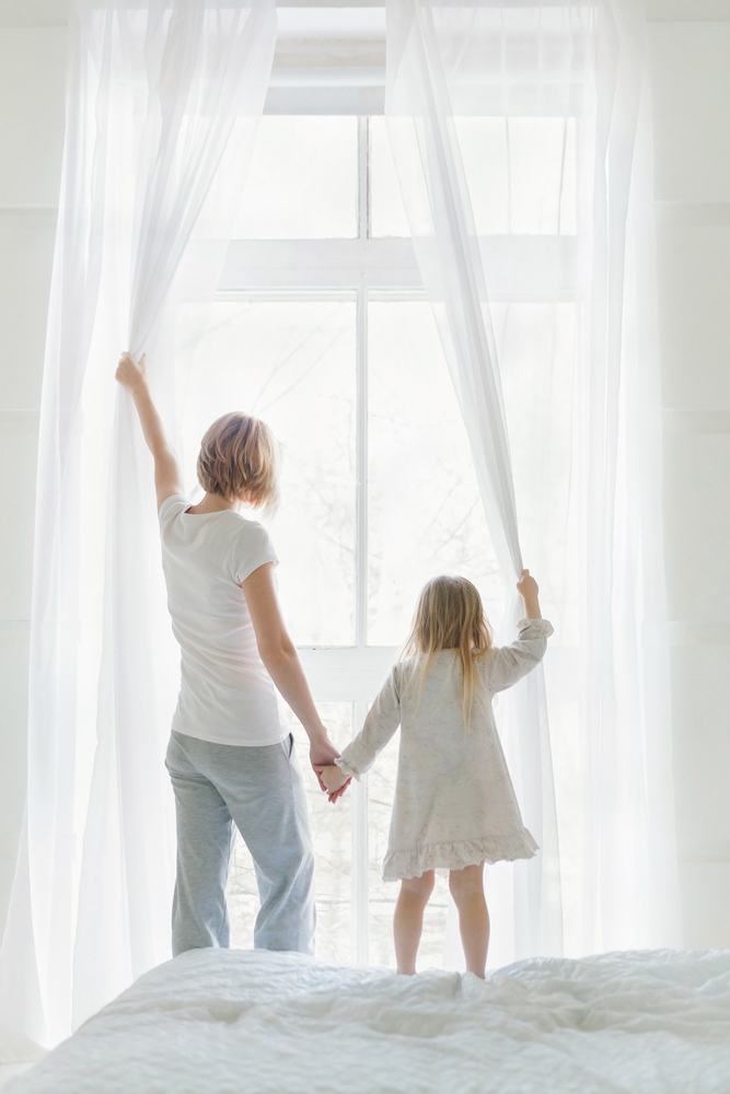 Young,Mother,With,Daughter,Opening,Window,Curtains,In,Cozy,Bedroom
